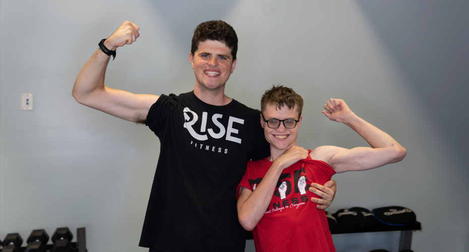 A coach and young male athlete are standing side by side, with their sleeves rolled up, smiling and flexing their biceps for the camera.